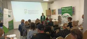 Pokaż zdjęcie: Widoczna prezentacja z logo PFRON a na jej tle jedna z organizatorek spotkania trzymająca w ręku mikrofon oraz widoczni pozostali uczestnicy