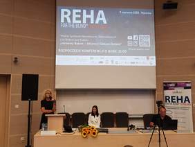 sala konferencyjna, przy stole siedzi kobieta i mężczyzna, za nimi ekran zawierający informacje o wydarzeniu, w szczególności duży napis reha oraz imię i nazwisko prelegenta, z lewej strony za mównicą kobieta