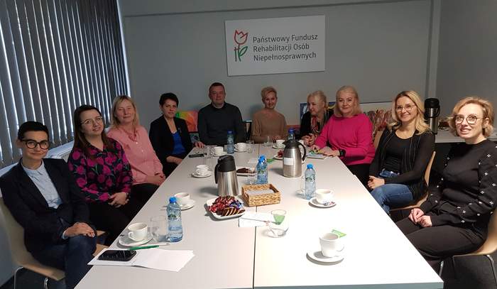 Pokaż zdjęcie: Przy białym, prostokątnym stole w sali konferencyjnej siedzi dziewięć osób z ośrodków edukacyjnych. Na ścianie za nimi wisi tablica z napisem „Państwowy Fundusz Rehabilitacji Osób Niepełnosprawnych”. Uczestnicy spotkania rozmawiają i wspólnie szukają sposobów, jak lepiej wspierać uczniów z niepełnosprawnościami.
