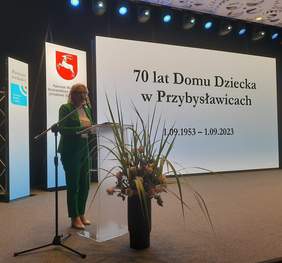 Pokaż zdjęcie: Na scenie przy mównicy stoi kobieta blondynka w zielonym garniurez. Przed mównicą stoi bukiet wysokich kwiatów, w tle prezentacja z hasłem 70-lecie Domu Dziecka w Przybysławicach