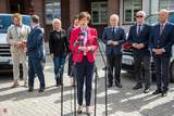 Konferencja prasowa przed budynkiem Starostwa Powiatowego w Pile. Na pierwszym planie kobieta o krótkich brązowych włosach przemawia do mikrofonów. Za nią stoi grupa siedmiu elegancko ubranych osób. Spotkanie dotyczy przekazania nowych busów dla Domów Pomocy Społecznej.