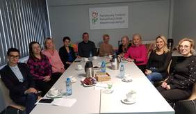 Pokaż zdjęcie: Przy białym, prostokątnym stole w sali konferencyjnej siedzi dziewięć osób z ośrodków edukacyjnych. Na ścianie za nimi wisi tablica z napisem „Państwowy Fundusz Rehabilitacji Osób Niepełnosprawnych”. Uczestnicy spotkania rozmawiają i wspólnie szukają sposobów, jak lepiej wspierać uczniów z niepełnosprawnościami.