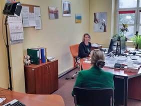 Pokaż zdjęcie: Przy biurku siedzą dwie panie. Jedna siedzi bokiem, a druga tyłem. W lewym górnym rogu widać reflektory światła. Na biurku leży stos dokumentów. 