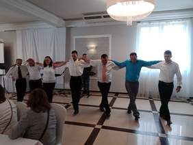 Pokaż zdjęcie: Na scenie grupa 7 osób w tym jedna kobieta trzyma się za ramiona, ustawiona w jednej linii,  tańczy grecki taniec sirtaki