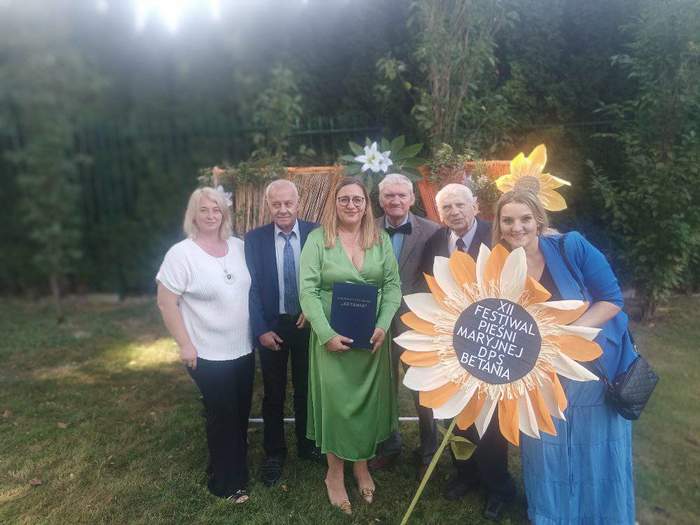 Pokaż zdjęcie: Zdjęcie grupy starszych osób, jedna z kobiet trzyma w ręku dekorację, kwiat rumianku z napisem XII Festiwal Pieśni Maryjnej.