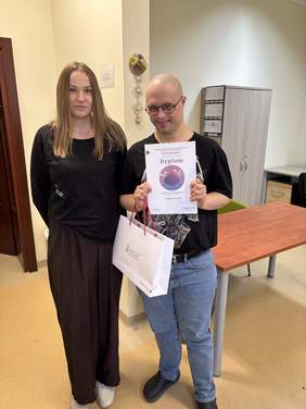 Pokaż zdjęcie: Kobieta stoi obok mężczyzny trzymającego dyplom i torbę z upominkiem. Oboje pozują do zdjęcia w pomieszczeniu z czerwonym stołem.