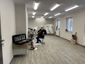 Pokaż zdjęcie: Na krześle siedzi mężczyzna ubrany na ciemno. W ręku trzyma gitarę. W jasnej sali z drewnianą podłogą znajdują się instrumenty muzyczne.