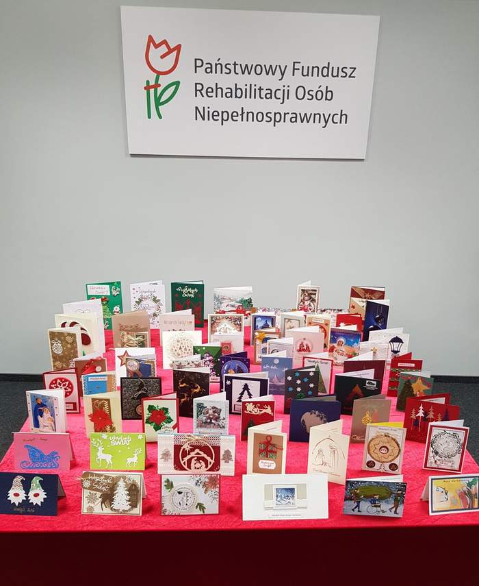 Pokaż zdjęcie: Kilkadziesiąt kartek bożonarodzeniowych ułożonych na stole przykrytym czerwonym płótnem. W tle widoczny jest przymocowany do ściany duży logotyp Państwowego Funduszu Rehabilitacji Osób Niepełnosprawnych.