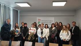 Pokaż zdjęcie: Grupa kilkunastu osób stoi w sali konferencyjnej na tle dużego logotypu PFRON zawieszonego na ścianie. Z przodu znajduje się rząd sześciu krzeseł. Okna przysłonięte są żaluzjami. Na suficie podwieszone są dwie lampy, które w danej chwili są włączone.