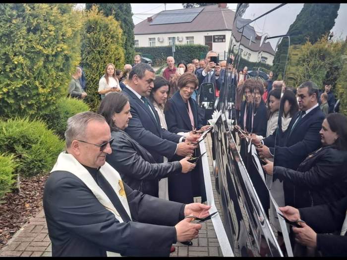 Pokaż zdjęcie: Grupa pięciu osób stoi bezpośrednio przy autobusie w kolorze czarnym i przecina symboliczną wstęgę. W tle kolejna grupa kilkunastu osób oraz budynek świetlicy wiejskiej.