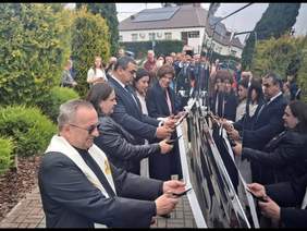 Pokaż zdjęcie: Grupa pięciu osób stoi bezpośrednio przy autobusie w kolorze czarnym i przecina symboliczną wstęgę. W tle kolejna grupa kilkunastu osób oraz budynek świetlicy wiejskiej.