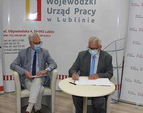 Pokaż zdjęcie: Z prawej strony mężczyzna w szarym garniturze siedzący przy okrągłym stoliku podpisuje dokumenty. Po lewej stronie siedzi mężczyzna w białym fotelu, ubrany w szary garnitur, trzyma na kolanach dokumenty. Za nimi ścianka z logo Wojewódzkiego Urz