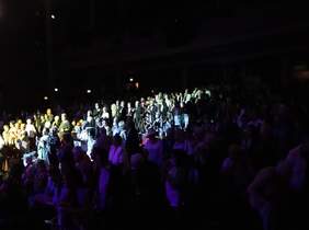 Pokaż zdjęcie: sala koncertowa w teatrze, zgaszone światło, po rodku widać widownię która stoi i oklaskuje wykonawców koncertu 