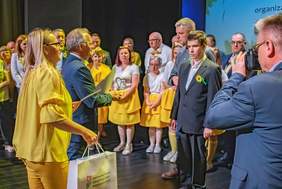 Pokaż zdjęcie: Oświetlona scena. Na scenie stoją laureaci ubrani w kolor żółty. Organizatorzy przekazują laureatom upominki z logo PFRON.