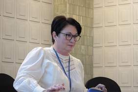 Pokaż zdjęcie: Kobieta z czarnymi krótkimi włosami i białej bluzce siedzi przy stole prezydialnym, przygotowuje się do konferencji