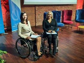 Pokaż zdjęcie: W sali znajdują się dwie panie siedzące na wózkach inwalidzkich. Prowadzą konferencję i uśmiechają się. Są to: Anna Płoszyńska absolwentka Uniwersytetu Medycznego w Łodzi, II Wicemiss Świata na Wózku 2022 oraz Monika Mazur z Fundacji Aktywnej Rehabilitacji. 