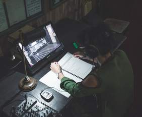 Pokaż zdjęcie: Fragment ciemnego pomieszczenia, kobieta ze słuchawkami na uszach siedzi i czyta tekst przedstawienia przed nią stoi monitor z podglądem na scenę teatralną.