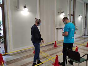 Pokaż zdjęcie: w pomieszczeniu na podłodze ułożony jest tor z żółtych drążków połączonych czerwonymi pachołkami przechodzi w nim kobieta z zasłoniętymi oczami i uszami z białą laską w dłoni obok asystuje mężczyzna w błękitnej koszulce 