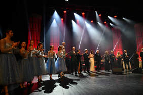 Kobiety i mężczyźni na scenie, kilka kobiet w niebieskich jednakowych sukienkach, pozostali w strojach wieczorowych