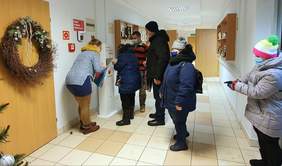 Pokaż zdjęcie: Długi jasny korytarz 5 osób stoi w kolejce do wysokiego urządzenia dozującego płyn do dezynfekcji rąk obok stoi kobieta i udziela instrukcji obsługi