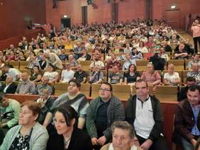 Pokaż zdjęcie: Na widowni siedzi bardzo dużo osób. Wszystkie miejsca są zajęte. Widzowie patrzą na scenę. 