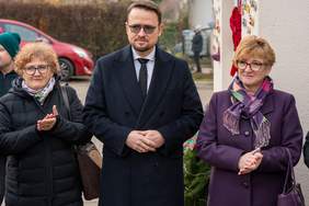 Pokaż zdjęcie: Trzy osoby stoją na zewnątrz. W centrum stoi mężczyzna w ciemnym płaszczu i okularach. Po jego lewej stronie (po prawej stronie zdjęcia) stoi kobieta w fioletowym płaszczu i szalu, klaszcząca w dłonie. Po jego prawej stronie (po lewej stronie zdjęcia) stoi inna kobieta w czarnej puchowej kurtce, również klaszcząca. W tle widać czerwony samochód.