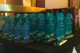 Pokaż zdjęcie: Zdjęcie przedstawia szereg szklanych statuetek w kolorze turkusowym, ustawionych na czarnym stole. Każda statuetka ma wygrawerowane logo oraz napisy, w tym nazwiska zwycięzców, numery miejsc (np. "MIEJSCE VII", "MIEJSCE IX", "MIEJSCE X") oraz dedykacje z podziękowaniami. Statuetki posiadają solidną, szklaną podstawę. W tle widać torby z upominkami lub nagrodami. 