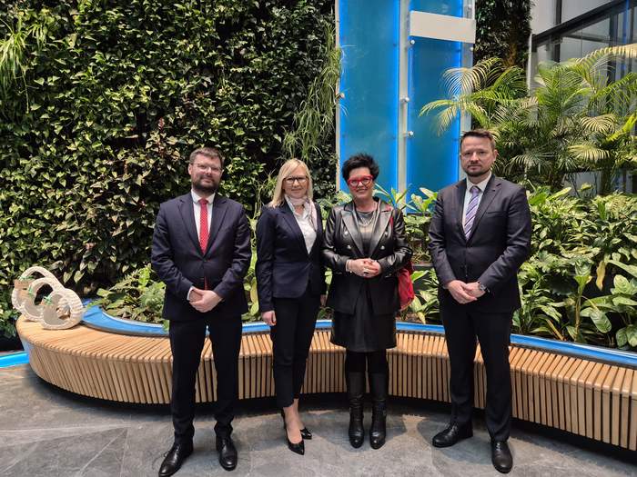 Pokaż zdjęcie: Czworo ludzi w eleganckich strojach pozuje do zdjęcia w jasnym, nowoczesnym wnętrzu pełnym zieleni. Mężczyźni mają na sobie ciemne garnitury, a kobiety – granatowy garnitur oraz czarną, skórzaną stylizację. W tle znajduje się ściana pokryta roślinami oraz niebieska, podświetlana instalacja.