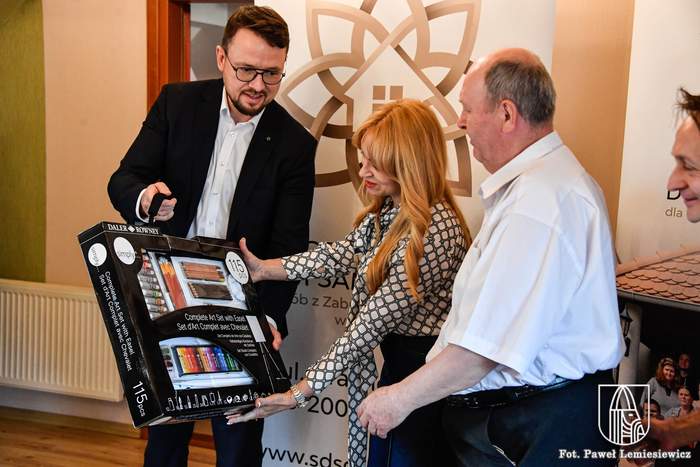 Pokaż zdjęcie: Dyrektor Oddziału wręcza laureatowi nagrodę, zestaw materiałów plastycznych w czarnym opakowaniu. 