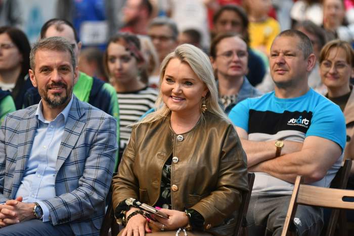 Pokaż zdjęcie: W centralnym punkcie uśmiechnięta kobieta, która zasiada na widowni, a obok niej inne osoby