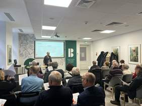 Pokaż zdjęcie: W sali wykładowej trwa prelekcja. Prelegentka stoi i omawia prezentację. Na widowni siedzą ludzie na krzesełkach i słuchają wystąpienia. 