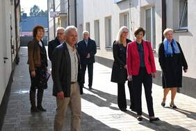 Pokaż zdjęcie: Grupa siedmiu osób znajduje się pomiędzy dwoma budynkami i porusza się chodnikiem wyłożonym nową kostką brukową.