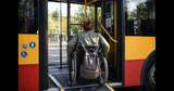 Do autobusu wjeżdża kobieta siedząca na wózku inwalidzkim. Na oparciu wózka inwalidzkiego przewieszony jest plecak. W tle widać wnętrze autobusu. 