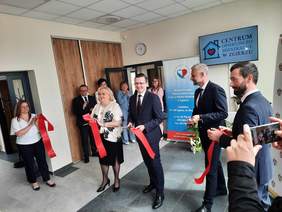 Pokaż zdjęcie: W pomieszczeniu odbyło się oficjalne przecięcie wstęgi. Niektóre osoby trzymają w dłoniach czerwoną szarfę, a inne - klaszczą. Na ścianie wisi tablica z nazwą: Centrum Opiekuńczo-Mieszkalne w Zgierzu. 