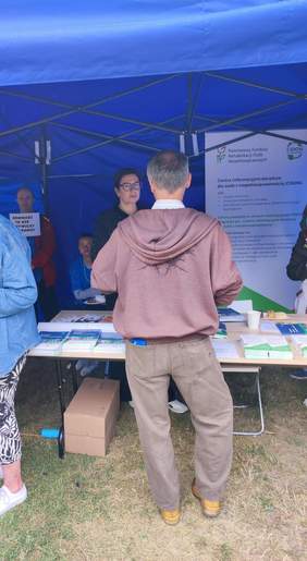 Pokaż zdjęcie: Mężczyzna w brązowej bluzie stoi tyłem przy stole informacyjnym pod niebieskim namiotem. Po drugiej stronie stołu znajduje się kobieta, która z nim rozmawia. W tle widoczny jest roll-up z logo PFRON i CIDON, a na stole leżą ulotki i broszury.