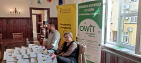 Pokaż zdjęcie: Kobieta z mężczyną siedzą przy stoliku wypełnionym materiałami informacyjnymi, a za nimi rollupy z logo PFRON i OWiT