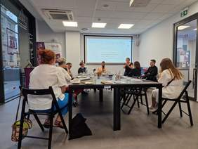 Pokaż zdjęcie: Grupa osób uczestniczy w warsztatach. Siedzą wokół stołu. Z przodu ekran rzutnikiem. Widać tłumaczy PJM.