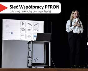 Pokaż zdjęcie: Scena konferencyjna z czarnym tłem i banerem „Sieć Współpracy PFRON”. Pod banerem widać ekran z prezentacją. Na prezentacji obrazy technologii asystujących i wspomagających dla osób z niepełnosprawnościami. Na środku zdjęcia ustawiony jest pulpit z otwartym laptopem. Po prawej stronie stoi kobieta o jasnej karnacji, w okularach, z długimi blond włosami, ubrana w białą koszulę i czarne spodnie. Trzyma w rękach mikrofon i jest skierowana w stronę publiczności. W górnej lewej części zdjęcia widoczny jest napis: „Sieć Współpracy PFRON” oraz mniejszy: „działamy razem, by pomagać lepiej”.