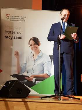 Mężczyzna w garniturze stoi na scenie przed mikrofonem, przemawia. Za nim duży baner na którym widać kobietę na wózku inwalidzkim