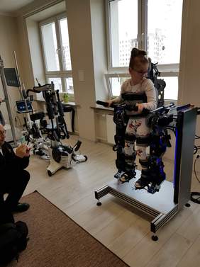 Pokaż zdjęcie: w pomieszczeniu Fundacji umieszczono nowe urządzenie o nazwie Prodrobot na którym stoi dziewczynka z niepełnosprawnością ruchową, nowe urządzenie pionizuje podopieczną Fundacji 