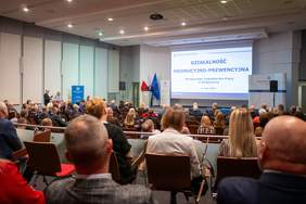 W sali konferencyjnej siedzą licznie zgromadzeni uczestnicy Gali podsumowujacej konkursy i programy Okręgowego Inspektoratu Pracy w Bydgoszczy. Słuchają przemawiającego prelegenta, który stoi przy mównicy.  Za nim widoczne są flagi Polski i Unii Europejskiej. W tle widoczny duży ekran z wyświetlaną prezentacją”