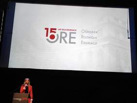 Kobieta za mównicą, z mikrofonem w ręce, w czerwonej marynarce, w tle napis 15 ORE na białym tle