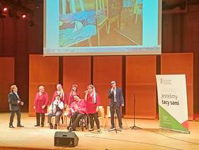 Pokaż zdjęcie: na scenie sali koncertowej siedzą i stoją wokaliści ze Studio Integracji kobiety są ubrane na czerwono i mają na głowie kolorowe wianki a mężczyżni na głowie mają kaszkiety. w rręku trzymają mikrofony i śpiewają w tle telebim na którym wy