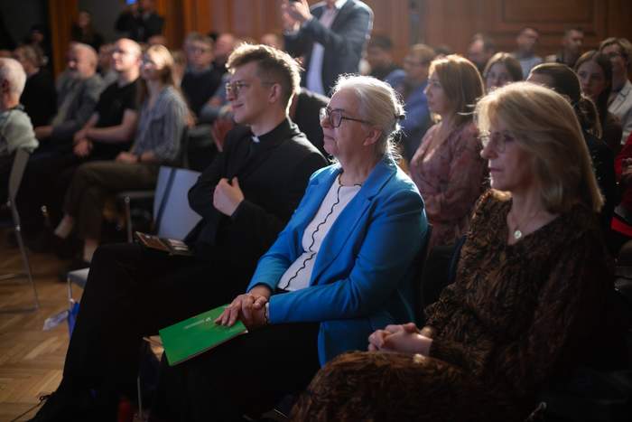 Pokaż zdjęcie: W pierwszym rzędzie sali konferencyjnej siedzą dwie kobiety oraz ksiądz. W tle rozmazane sylwetki kilkudziesięciu osób znajdujących się na sali.