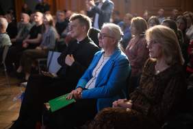 Pokaż zdjęcie: W pierwszym rzędzie sali konferencyjnej siedzą dwie kobiety oraz ksiądz. W tle rozmazane sylwetki kilkudziesięciu osób znajdujących się na sali.