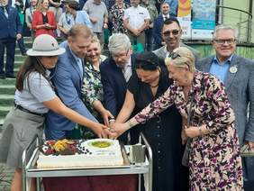 Pokaż zdjęcie: Na stole leży duży, prostokątny tort. Nad nim pochylaj się ludzie. Wspólnie trzymają w rękach nóż i próbują ukroić pierwszy kawałek tortu. 