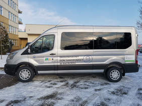 Pokaż zdjęcie: Na parkingu przed budynkiem stoi ustawiony bokiem dziewięcioosobowy srebrny bus. Na drzwiach od strony kierowcy ma zamieszczone logo powiatu raciborskiego, na środkowych duży nas środowiskowy dom samopomocy w Raciborzu, z tyłu logo PFRON.