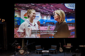 Pokaż zdjęcie: Na scenie zawieszony jest ogromny ekran. Wyświetlona jest na nim rozmowa dwóch kobiet. Pod ekranem stoją instrumenty muzyczne, fotele oraz stolik. 