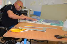 Pokaż zdjęcie: Mężczyzna na wózku inwalidzkim i w okularach pochylający się nad stołem i pracujący nad tablicą graficzną informacji o szpitalu