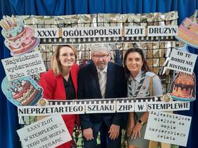 Pokaż zdjęcie: Przed ścianką do zdjęć stoją trzy osoby: dwie panie i jeden pan. Nad ich głowami znajduje sie napis: XXXV Ogólnopolski Zlot Drużyn Nieprzetartego Szlaku ZHP. Wokół nich są kolorowe dekoracje. 